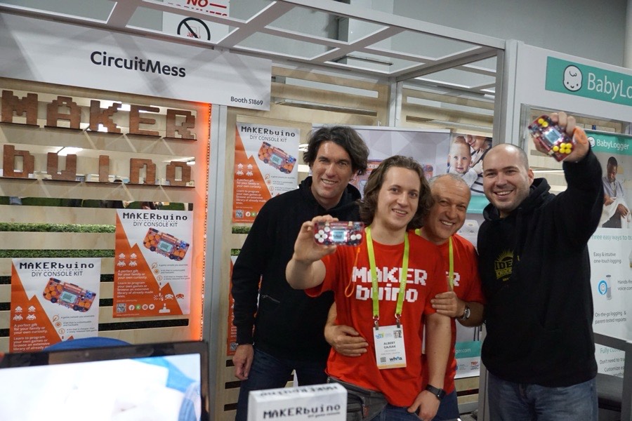 Albert Gajšak at a MAKERbuino trade show booth with three visitors, all holding up consoles.