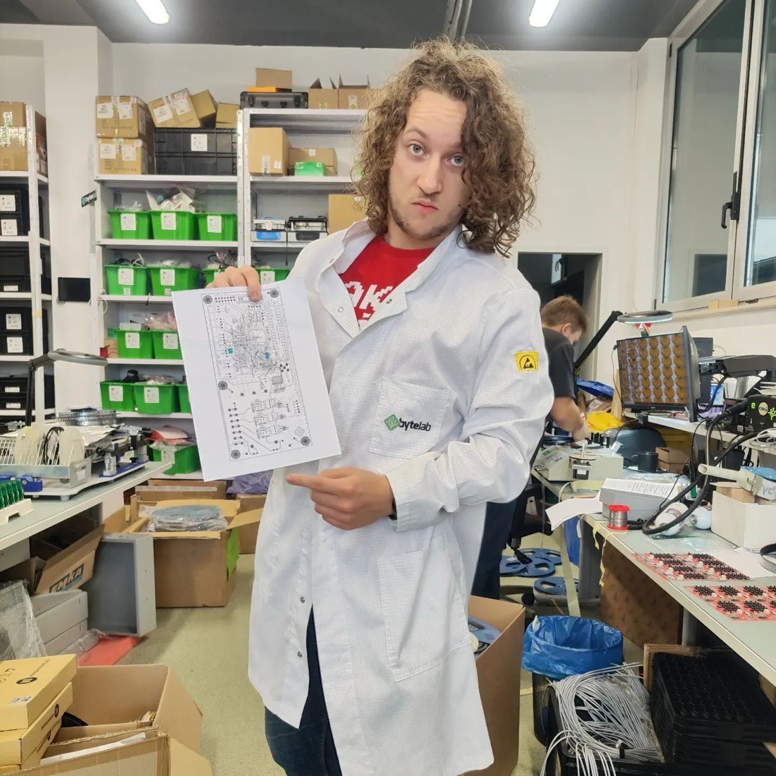 Albert Gajšak in a lab coat on the production floor, holding a printout of a circuit board layout.