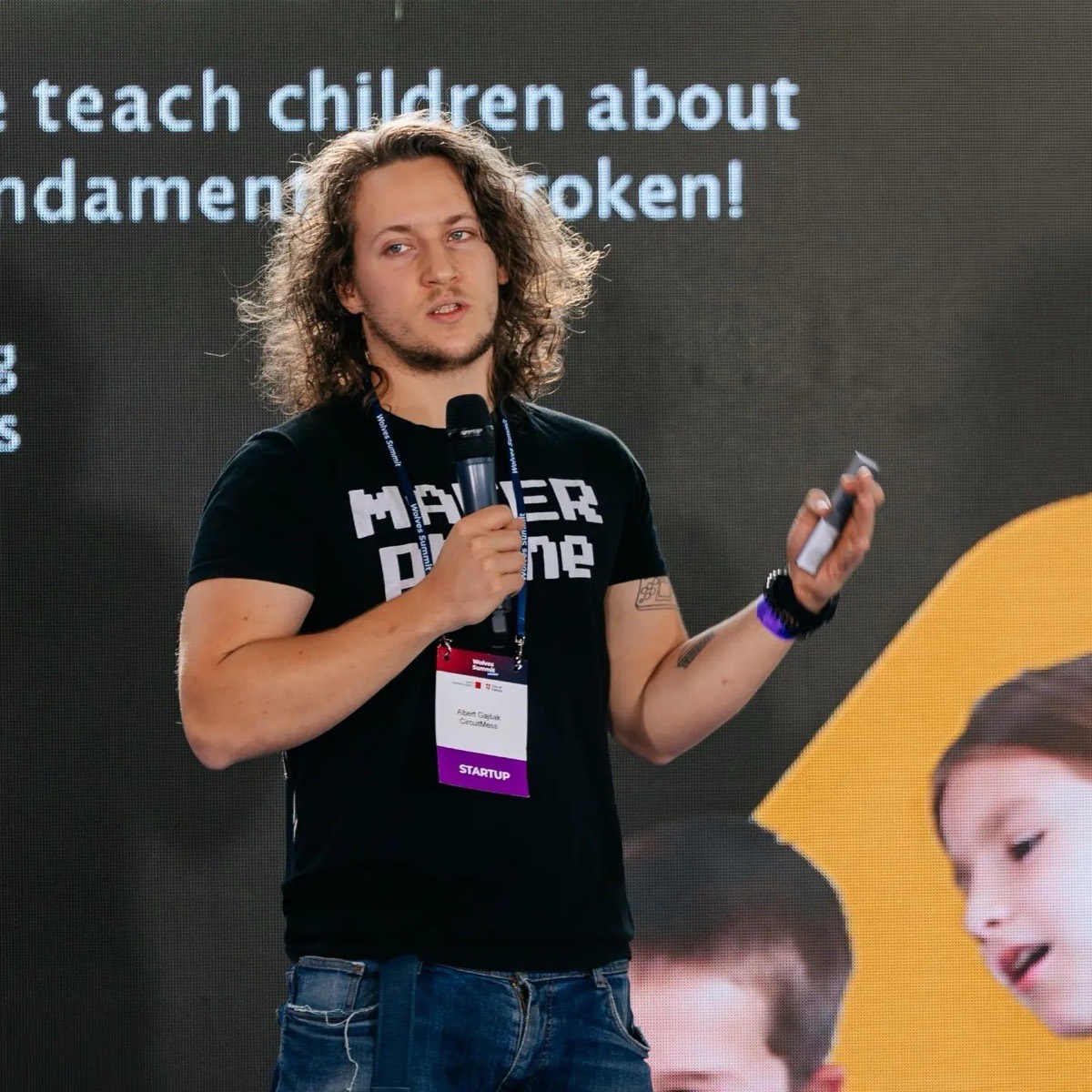 Albert Gajšak on stage with a microphone in a MAKERphone T-shirt, in front of a slide reading “we teach children about … broken!”