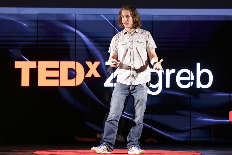 Albert Gajšak speaking on the TEDxZagreb stage in front of a large illuminated TEDxZagreb sign.