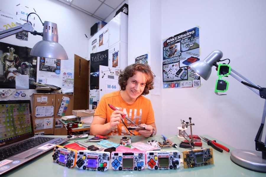 Teenage Albert Gajšak at a cluttered workshop desk with a row of five finished handheld consoles in different colours.