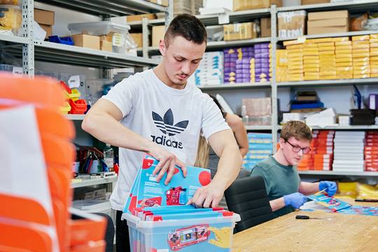 Two CircuitMess staff in the stockroom lifting flat-packed Bit cartons out of a tub, shelves of boxed product behind them.