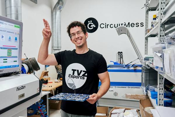 A CircuitMess employee holding out a freshly populated panel of circuit boards, with the pick-and-place machine behind them.