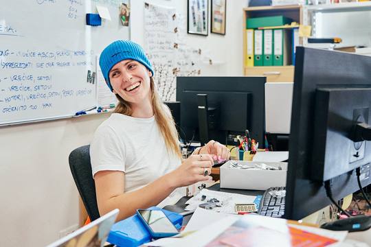 A CircuitMess employee in a blue beanie laughing over a tray of components, a whiteboard of Croatian production notes behind her.