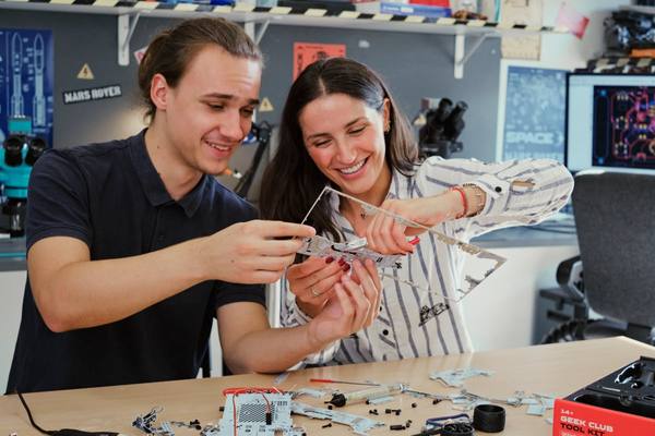 Two colleagues laughing as they fit together a clear acrylic rover frame, a Mars rover poster on the wall behind.