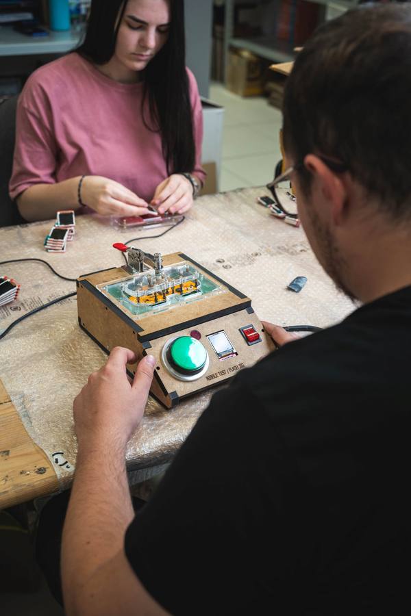 A hand pressing the green button on a plywood test-and-flash jig, while a colleague opposite hand-assembles Nibble consoles.