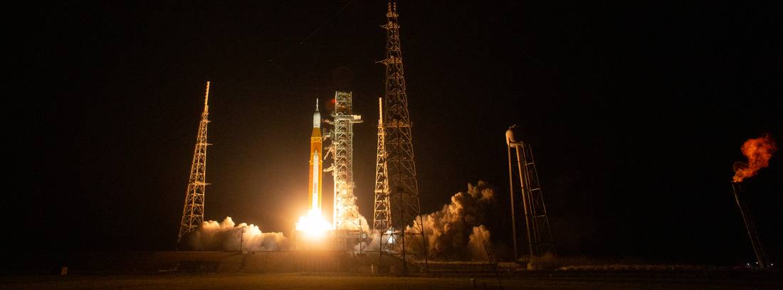 Artemis I lifting off at night against a black sky, the rocket plume lighting the launch tower.